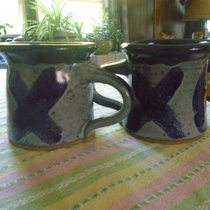 May include: Two handmade ceramic mugs. Each mug has a speckled gray base with a dark blue X design. The rims and handles are dark blue. The mugs sit on a striped tablecloth.