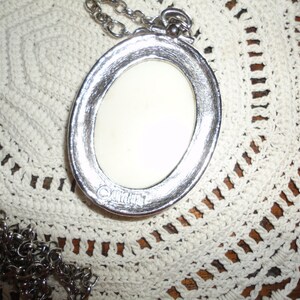 May include: A silver oval pendant necklace with a white cameo. The cameo is set in a silver frame and has a raised design. The necklace is on a silver chain.
