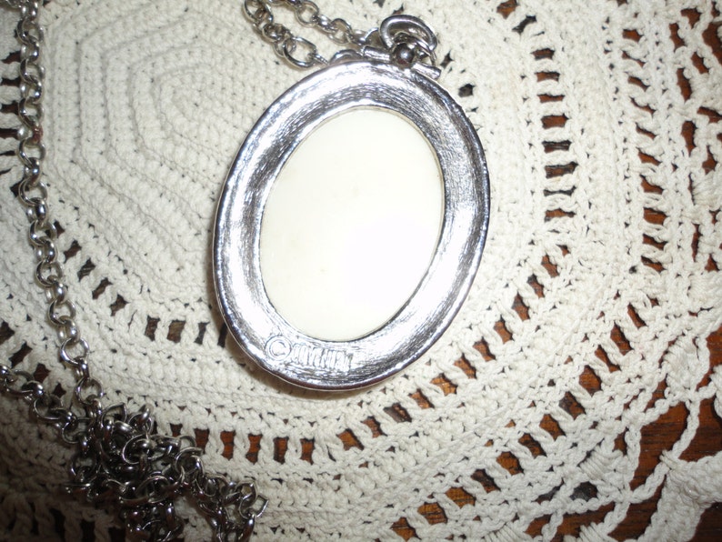 May include: A silver oval pendant necklace with a white cameo. The cameo is set in a silver frame and has a raised design. The necklace is on a silver chain.