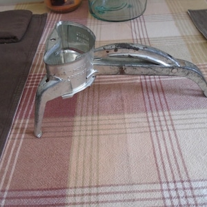 May include: A vintage, silver-colored citrus juicer with a lever and a cup-shaped receptacle. The juicer has a sturdy base with three legs and is designed for manual operation. The metal shows signs of age and use.