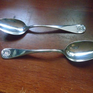 May include: Two silver spoons with a reflective surface are displayed on a wooden surface. Each spoon has an oval bowl and a handle with a decorative floral design at the end. The spoons are positioned parallel to each other.