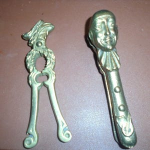 May include: Two antique brass nutcrackers. One is a traditional design with a leaf motif, the other is a whimsical design with a face.