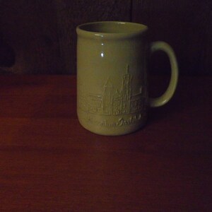 May include: A beige ceramic mug with a raised design of a city skyline and the text "Westfalia Ostbild".