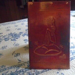 May include: A rectangular copper wall plaque with a dark, oxidized patina. The surface features an embossed design of a small house sitting on a hill, with three small metal grommets along the top edge for mounting purposes.