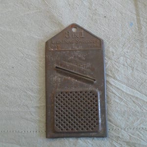 May include: A vintage 3-in-1 Lightning Shredder, made of brown metal. The kitchen tool has a rectangular shape with a perforated grating area and a slicing blade. The text "3 in 1 Lightning Shredder" is visible.