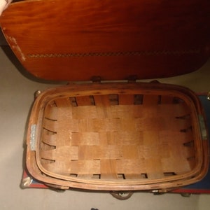 Puede incluir: Una cesta de picnic de madera con tapa abatible. La cesta est&aacute; hecha de madera trenzada y tiene un tinte marr&oacute;n. La tapa es de madera y tiene un tinte marr&oacute;n claro. La cesta est&aacute; vac&iacute;a.