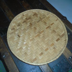 May include: A round, woven bamboo tray with a herringbone pattern. The tray is light brown and sits on a dark wooden surface. The tray's rim is also made of woven bamboo.