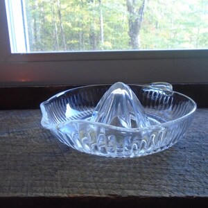May include: A clear glass citrus juicer with a ridged design. The juicer has a central cone with a pointed top and a wide, shallow base.