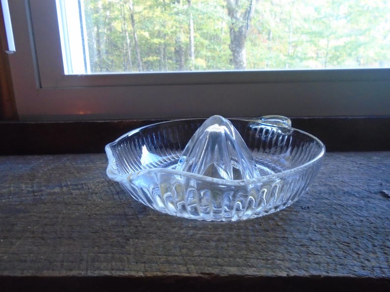 May include: A clear glass citrus juicer with a ridged design. The juicer has a central cone with a pointed top and a wide, shallow base.
