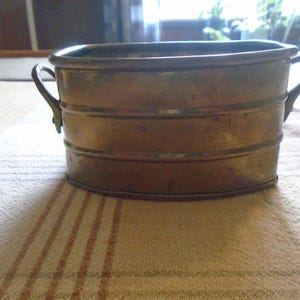 May include: A small, antique brass bucket with two curved handles. The bucket has a rectangular shape with rounded corners and horizontal bands around the body. The brass has a warm, aged patina, suggesting its vintage. This decorative piece could be used for storage or display.