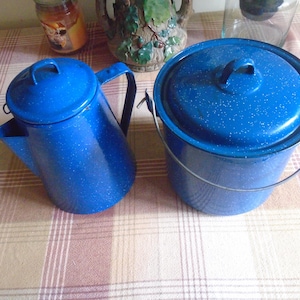 May include: Two blue enamelware camping items: a coffee pot with a lid and handle, and a pot with a lid and a wire handle. Both are speckled with white and sit on a plaid surface. The coffee pot has a spout.