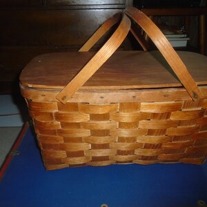 Puede incluir: Una cesta de picnic de madera vintage con un dise&ntilde;o tejido y una tapa con bisagras. La cesta tiene dos asas de madera y est&aacute; cerrada.