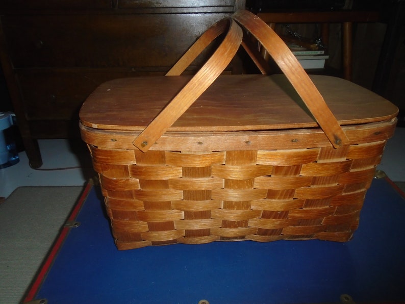 Puede incluir: Una cesta de picnic de madera vintage con un dise&ntilde;o tejido y una tapa con bisagras. La cesta tiene dos asas de madera y est&aacute; cerrada.