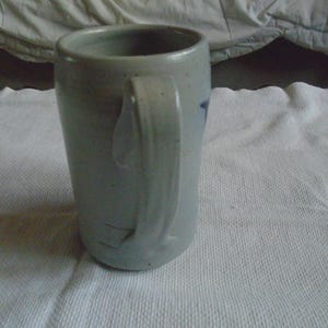 May include: A stoneware mug with a light gray glaze and a curved handle. The mug has a cylindrical shape and a slightly textured surface. A small blue design is visible on the side. The mug is sitting on a white surface.