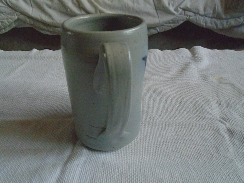 May include: A stoneware mug with a light gray glaze and a curved handle. The mug has a cylindrical shape and a slightly textured surface. A small blue design is visible on the side. The mug is sitting on a white surface.