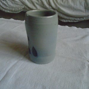 May include: A handmade, light gray ceramic tumbler with a cylindrical shape. The tumbler features dark purple leaf-shaped designs and a smooth, unglazed finish. The mug is suitable for hot or cold beverages.