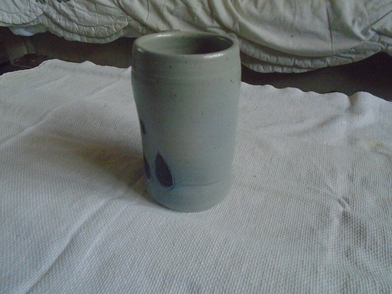May include: A handmade, light gray ceramic tumbler with a cylindrical shape. The tumbler features dark purple leaf-shaped designs and a smooth, unglazed finish. The mug is suitable for hot or cold beverages.