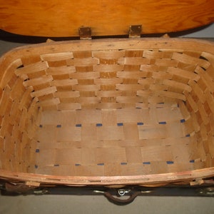Puede incluir: Una cesta de picnic de madera con tapa con bisagras y correas de cuero. La cesta est&aacute; hecha de madera tejida y tiene un forro a cuadros azul y blanco.