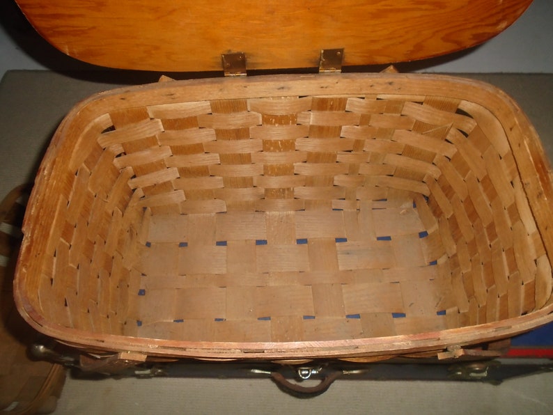Puede incluir: Una cesta de picnic de madera con tapa con bisagras y correas de cuero. La cesta est&aacute; hecha de madera tejida y tiene un forro a cuadros azul y blanco.