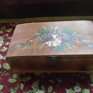 May include: A wooden box with a hinged lid and a painted floral design. The box has a brown stain and features a white flower with green leaves and pine cones.