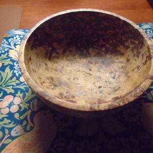May include: A rustic, wooden bowl with a natural, mottled finish. The bowl's interior and exterior display a warm, earthy color palette with brown and tan tones. The bowl sits on a blue floral patterned surface.
