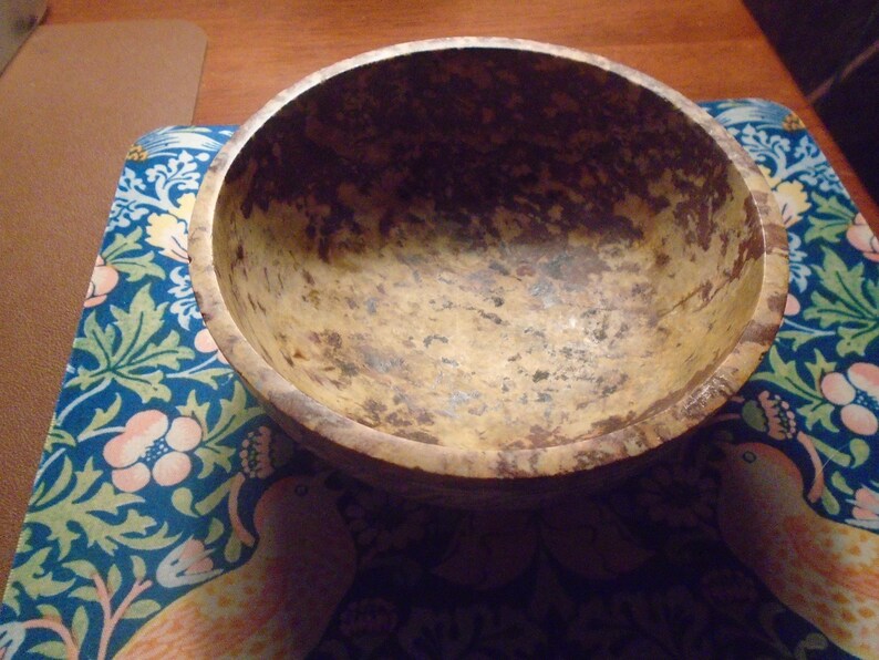 May include: A rustic, wooden bowl with a natural, mottled finish. The bowl's interior and exterior display a warm, earthy color palette with brown and tan tones. The bowl sits on a blue floral patterned surface.