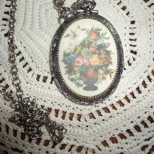 May include: A silver chain necklace with an oval pendant featuring a floral embroidery design. The pendant has a silver frame and a white background.
