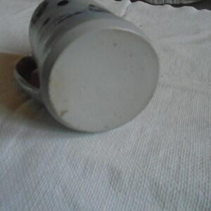 May include: A ceramic mug with a handle, resting on its side. The mug is primarily light gray with darker gray accents and a few dark spots near the top. The bottom of the mug is a lighter shade of gray. The mug is on a white surface.