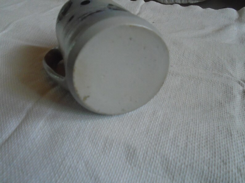 May include: A ceramic mug with a handle, resting on its side. The mug is primarily light gray with darker gray accents and a few dark spots near the top. The bottom of the mug is a lighter shade of gray. The mug is on a white surface.