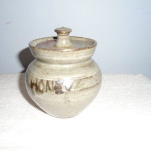 May include: A small, brown and white ceramic honey pot with a lid. The pot has a brown glaze and the word "Honey" is written on the side.