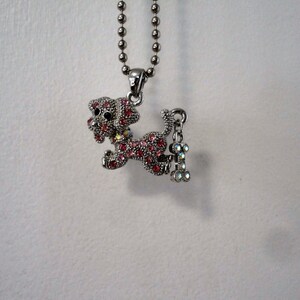 Pu&ograve; includere: Una collana d'argento con un ciondolo a forma di cane in cristallo rosa e un ciondolo a forma di osso. Il cane sta correndo e ha gli occhi neri.