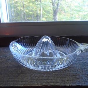 May include: A clear glass citrus juicer with a ribbed design and a handle. The juicer has a pointed center section for squeezing citrus fruits.