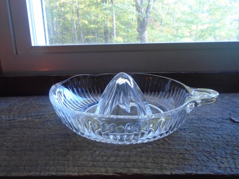 May include: A clear glass citrus juicer with a ribbed design and a handle. The juicer has a pointed center section for squeezing citrus fruits.