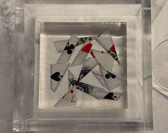 May include: A clear acrylic box containing a white napkin with several playing cards folded into triangles on top. The cards have various suits and numbers.