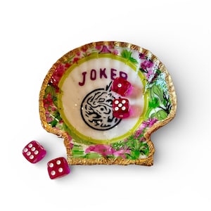 May include: A decorative, shell-shaped tray with a gold rim, floral accents, and the word "JOKER" in purple. Three red dice are inside the tray, and three more dice are scattered around it. The tray has a white center with a black design.