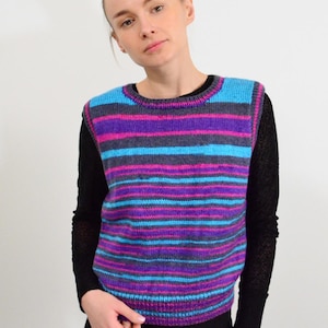 Può includere: Un maglione senza maniche a righe orizzontali nei toni del blu, viola e grigio. Il maglione è indossato sopra una camicia nera a maniche lunghe e pantaloni neri. Il design del maglione è un motivo colorato e accattivante.
