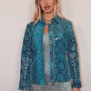 Op de afbeelding: Een jas met lange mouwen en een levendig turquoise en zwart slangenleermotief. De jas heeft een klassieke kraag, een zilverkleurige ritssluiting over de hele lengte aan de voorzijde en gestructureerde manchetten. Gedragen over een lichtgrijze jurk met bloemenprint.