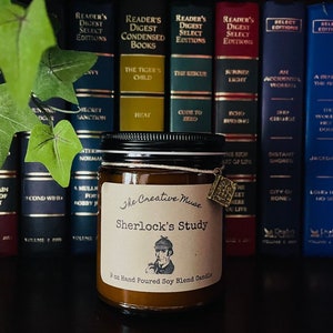May include: A brown glass candle jar with a black lid and a brown paper label. The label has the text "The Creative Muse" and "Sherlock's Study" in black lettering. The candle is 9 ounces and is a hand-poured soy blend candle. The candle is sitting on a shelf with a row of books behind it.