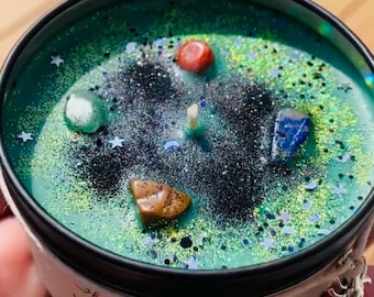 May include: A teal-colored candle in a black tin, adorned with gemstones and glitter. The candle is labeled "Witch's Brew" and has a silver charm attached. The candle's wax is decorated with stars and glitter.