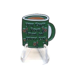 May include: A green enamel mug pin with the text "You can't pour from an empty cup." The mug has a white interior and is decorated with small orange flowers. The pin is displayed on a clear acrylic stand.