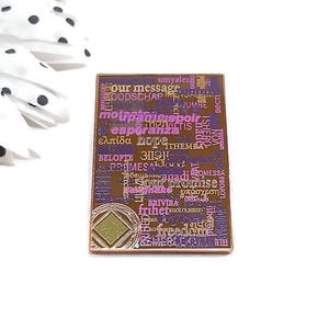 May include: A rectangular pin with a gold border and a purple and brown center. The center features words in various languages, including "hope" and "freedom." A geometric design is at the bottom.