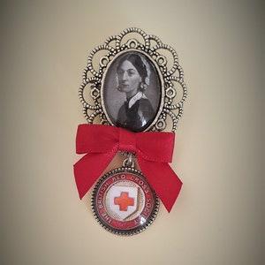May include: A silver brooch with a red bow and a round medallion featuring a red cross and the text "The British Red Cross Society". The medallion is attached to a cameo with a black and white image of a woman.