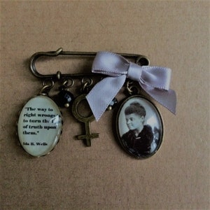 May include: A bronze charm bracelet featuring a quote by Ida B. Wells, a small portrait of a woman, a female symbol charm, and a lavender ribbon.