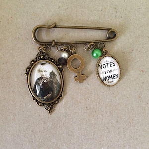 Puede incluir: Un broche de seguridad de bronce con abalorios, incluyendo un retrato de una mujer, un símbolo femenino y un abalorio que dice "Votes for Women".