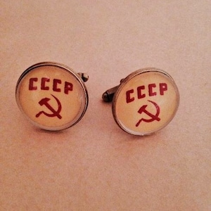 Op de afbeelding: Een paar zilveren manchetknopen met een witte achtergrond en een rood hamer-en-sikkel symbool. De manchetknopen hebben de tekst "CCCP" erop.