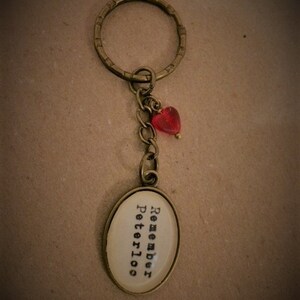 Könnte beinhalten: Ein Schlüsselanhänger aus Bronze mit einem runden Anhänger, auf dem in schwarzer Schrift "Remember Peterloo" steht. Ein kleiner roter Herzanhänger hängt an der Kette.