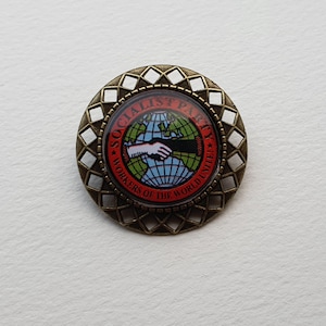 Pode incluir: Um broche redondo de cor bronze com um design branco e vermelho. O broche apresenta um globo com um aperto de mão e o texto "Socialist Party Workers of the World Unite".