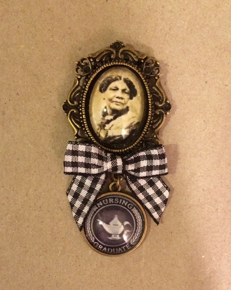 May include: A vintage brooch with a portrait in a bronze-toned frame. Below the frame is a black and white gingham bow, and a circular pendant with "NURSING GRADUATE" and a teapot design. The brooch has a classic, antique aesthetic.