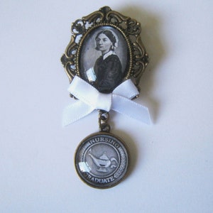 Pode incluir: Um broche de bronze com uma fita branca e um camafeu de vidro com uma mulher com um chapéu branco. O camafeu é rodeado por uma moldura de bronze ornamentada. O broche também apresenta um amuleto redondo de bronze com as palavras "Nursing Graduate" e uma imagem branca de uma lâmpada.