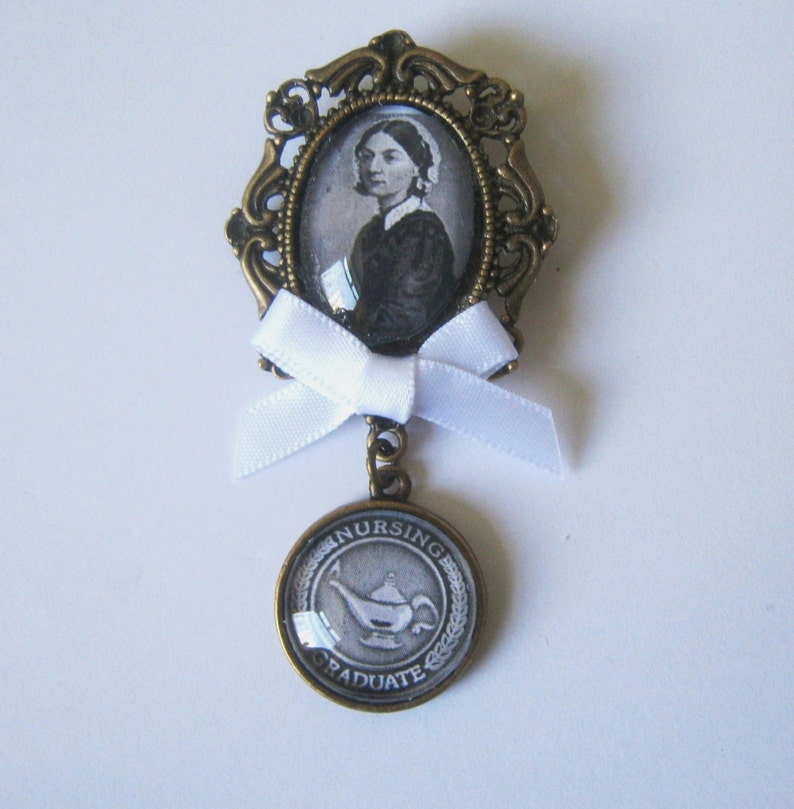 May include: A bronze pin with a white ribbon and a glass cameo featuring a woman in a white cap. The cameo is surrounded by an ornate bronze frame. The pin also features a round bronze charm with the words "Nursing Graduate" and a white image of a lamp.